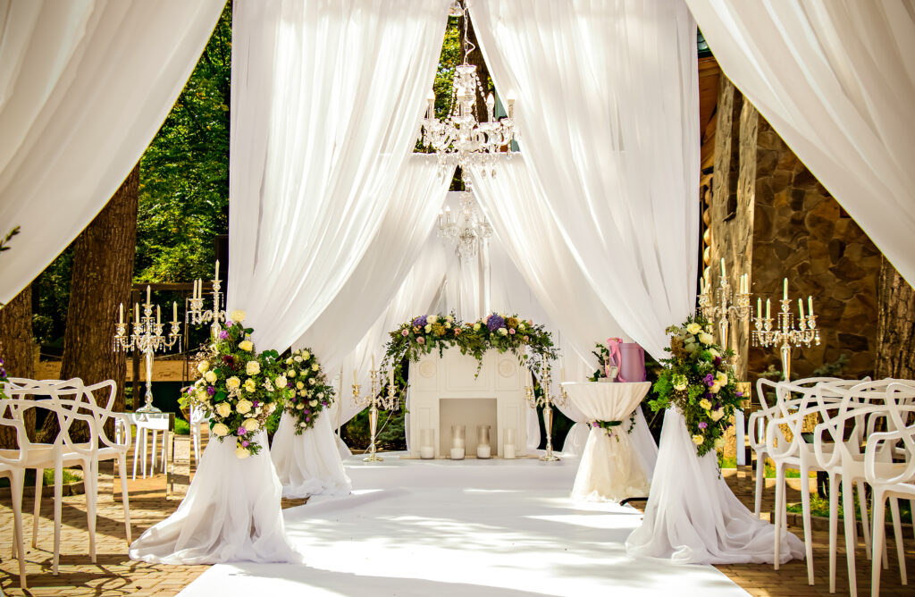 | Place for wedding ceremony in white color ,with white fireplace and chandeliers decorated with flowers and white cloth and wooden chairs for guests on each side outdoors.