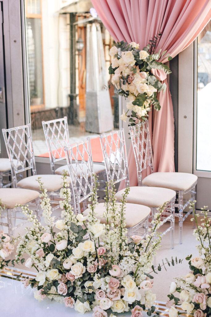| Rows of transparent chairs decorated with flower compositions of roses and white buttercups in the wedding ceremony area